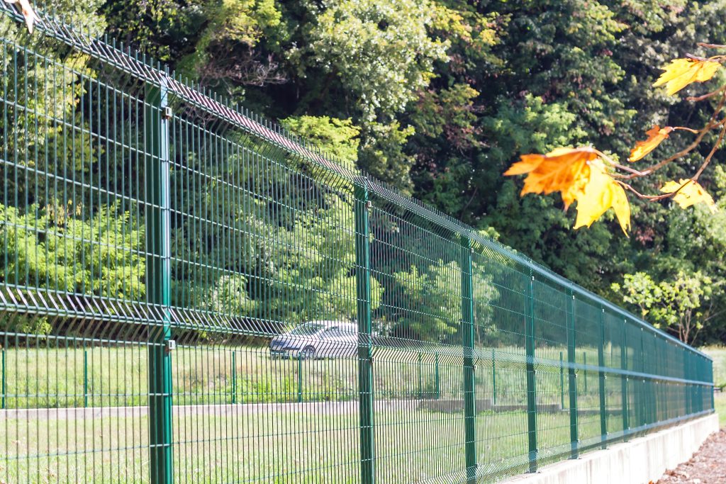 Green powder-coated welded mesh security fence panels with vertical steel posts, running alongside a paved path with trees in the background