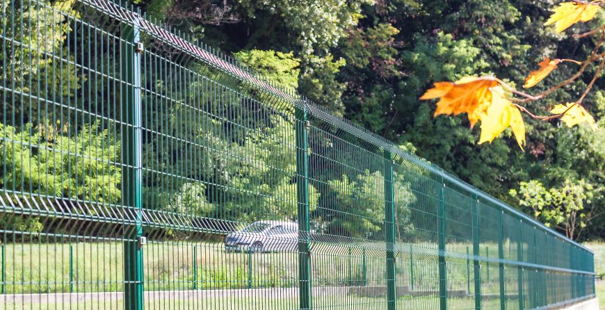 Green powder-coated welded mesh security fence panels with vertical steel posts, running alongside a paved path with trees in the background